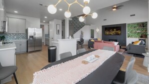 Cozinha moderna com ilha central, área de jantar com mesa grande e sala de estar com lareira e assentos.