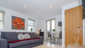 A modern living room with a grey sofa, a red throw blanket, and a white shag rug. There is a dining area with a table and chairs, and a wooden door with a glass panel. - Corner by the Canal - Leeds Central Duplex - Pass the Keys (Leeds)