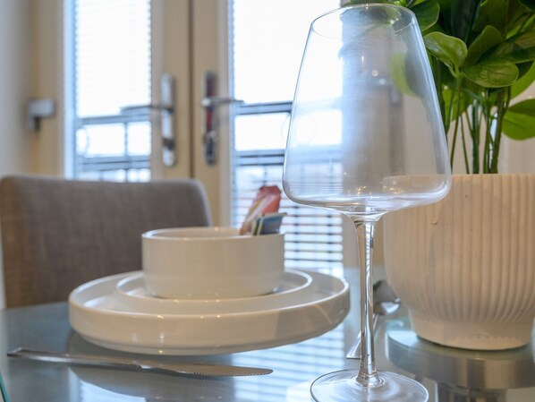 Una mesa de comedor con un plato blanco, un vaso y un cuchillo. En el fondo, hay un bol blanco y una planta en maceta.