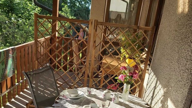 Une terrasse avec une table dressée pour deux personnes, entourée d’une clôture en treillis de bois, et une vue sur un bâtiment en arrière-plan.