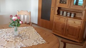Une table à manger en bois recouverte d’une nappe en dentelle, un vase rempli de fleurs, une chaise en bois avec un coussin rouge et un buffet en bois doté de portes en verre.