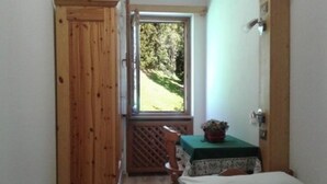 A room with a wooden door, a window with a view of trees, a small table with a green tablecloth, and a bed with a white pillow. - Cozy apartment with beautiful view of the Olympic Cortina d' Ampezzo (Cortina d'Ampezzo)