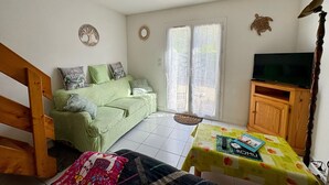 A room with a green sofa, a wooden cabinet, a television, and a table with a book and remote control. - Charming holiday home 400m from the beach (Saint-Hilaire-de-Riez)
