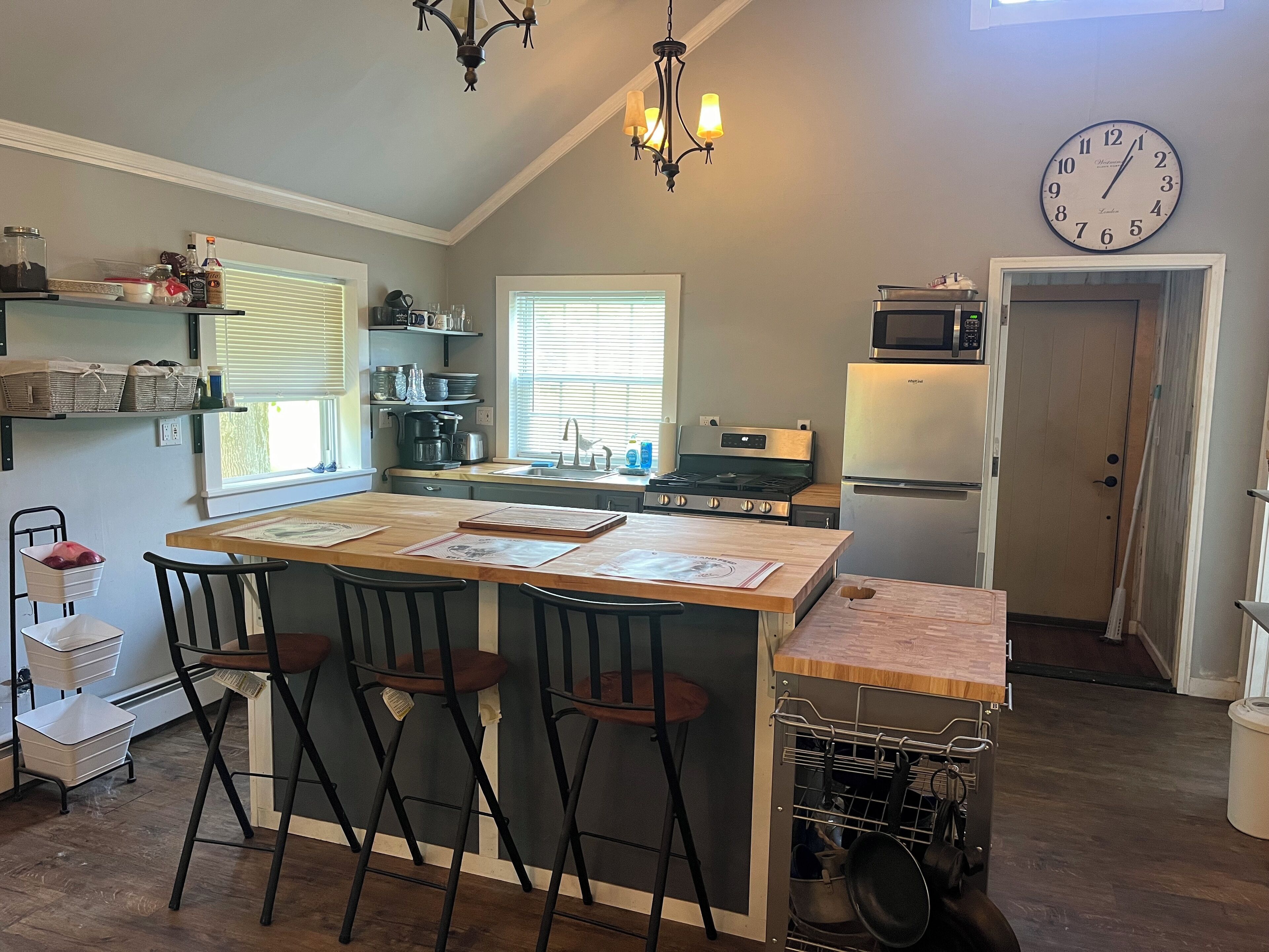 Una cocina con una isla central, taburetes de bar, microondas, refrigerador y un reloj en la pared.
