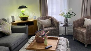 Un salón con un sofá gris, dos sillas de mimbre, una pequeña mesa de madera con una lámpara, una bandeja con un jarrón y un control remoto, y vistas al exterior a través de una ventana con cortinas.