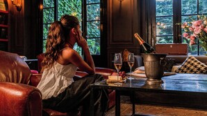 Una donna seduta su un divano in pelle in una stanza con vista su del verde attraverso ampie finestre, un tavolo con una bottiglia di champagne, calici e un vaso di fiori.
