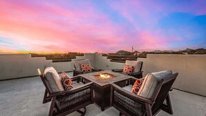A rooftop terrace with a fire pit table surrounded by wicker chairs, offering a view of the sunset over a cityscape. - Relaxing 3BR Haven w/ Pool & BBQ for Family Time (Mesa)