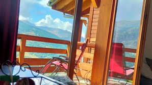Una habitación con vistas a un paisaje montañoso a través de amplios ventanales. Hay un atril de metal con frutas y un objeto negro.