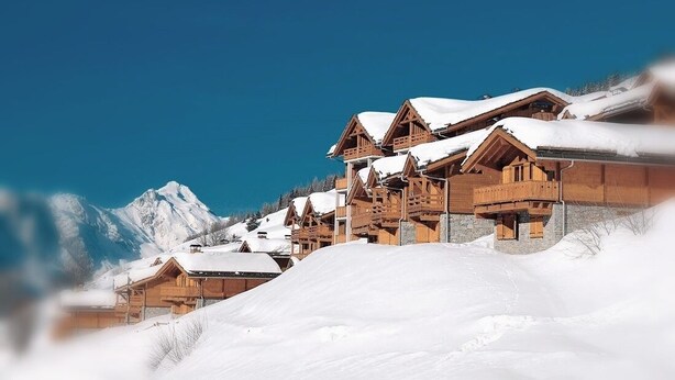 Un conjunto de cabañas de madera con tejados nevados, frente a un cielo azul despejado y una montaña.