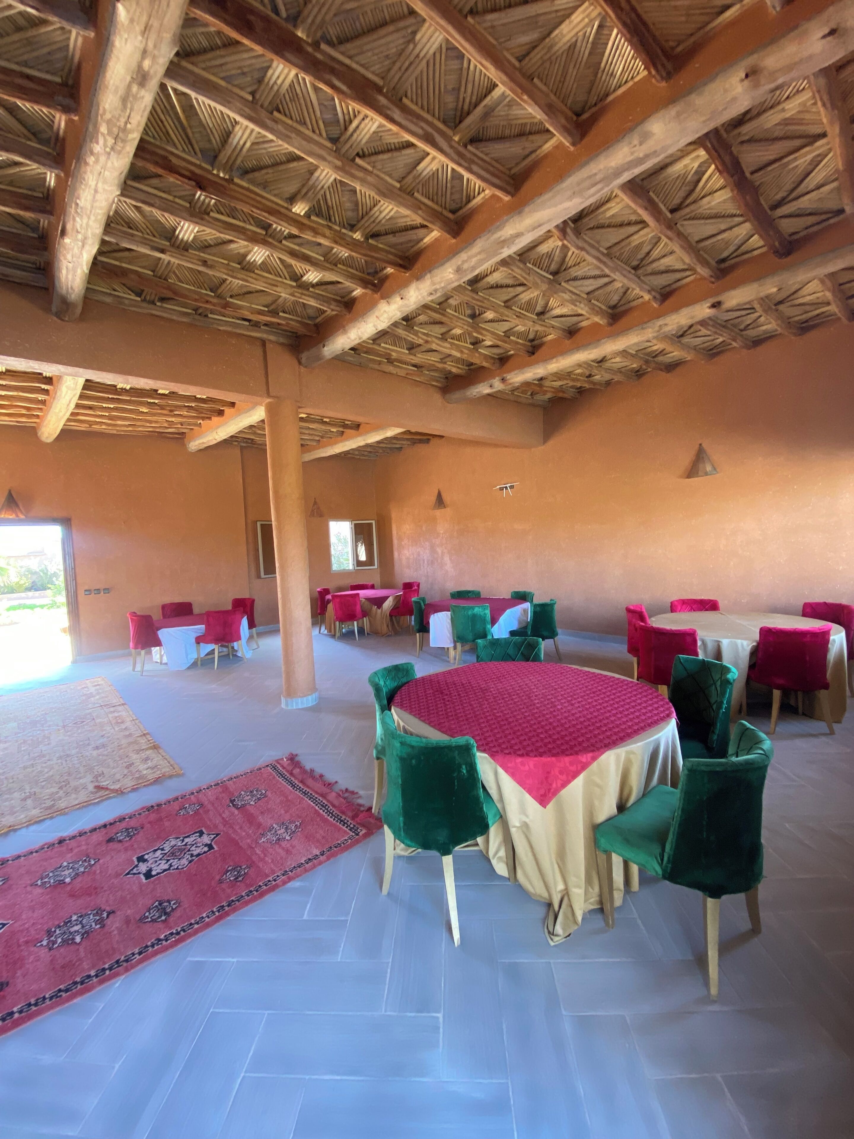 A spacious indoor area with multiple round tables and chairs, a patterned rug, and a wooden ceiling structure.