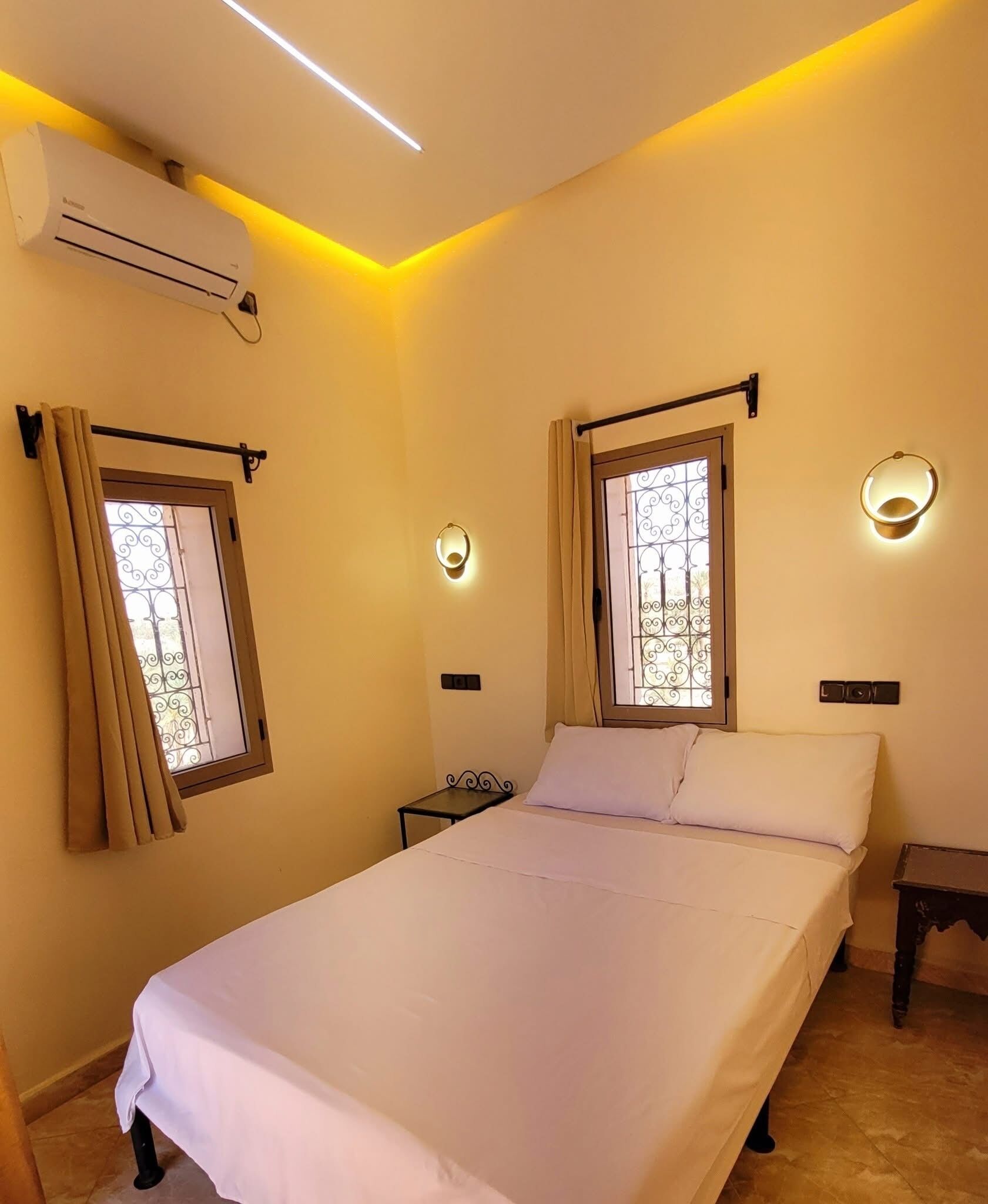 A bedroom with a bed, two windows with curtains, an air conditioner, a nightstand, and wall-mounted lights.