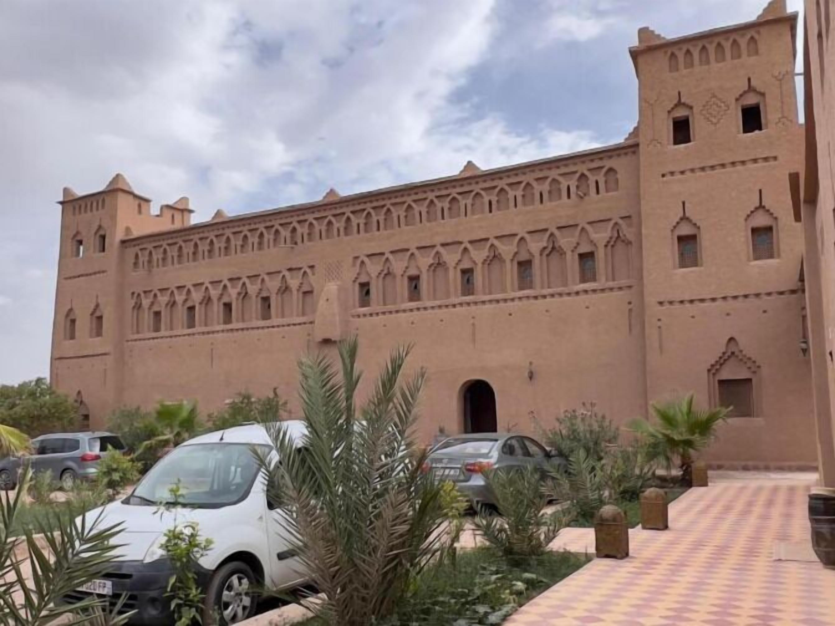 Un edificio grande de color beige con techo plano y varias ventanas de arco. Palmeras y un camino a cuadros en el frente.