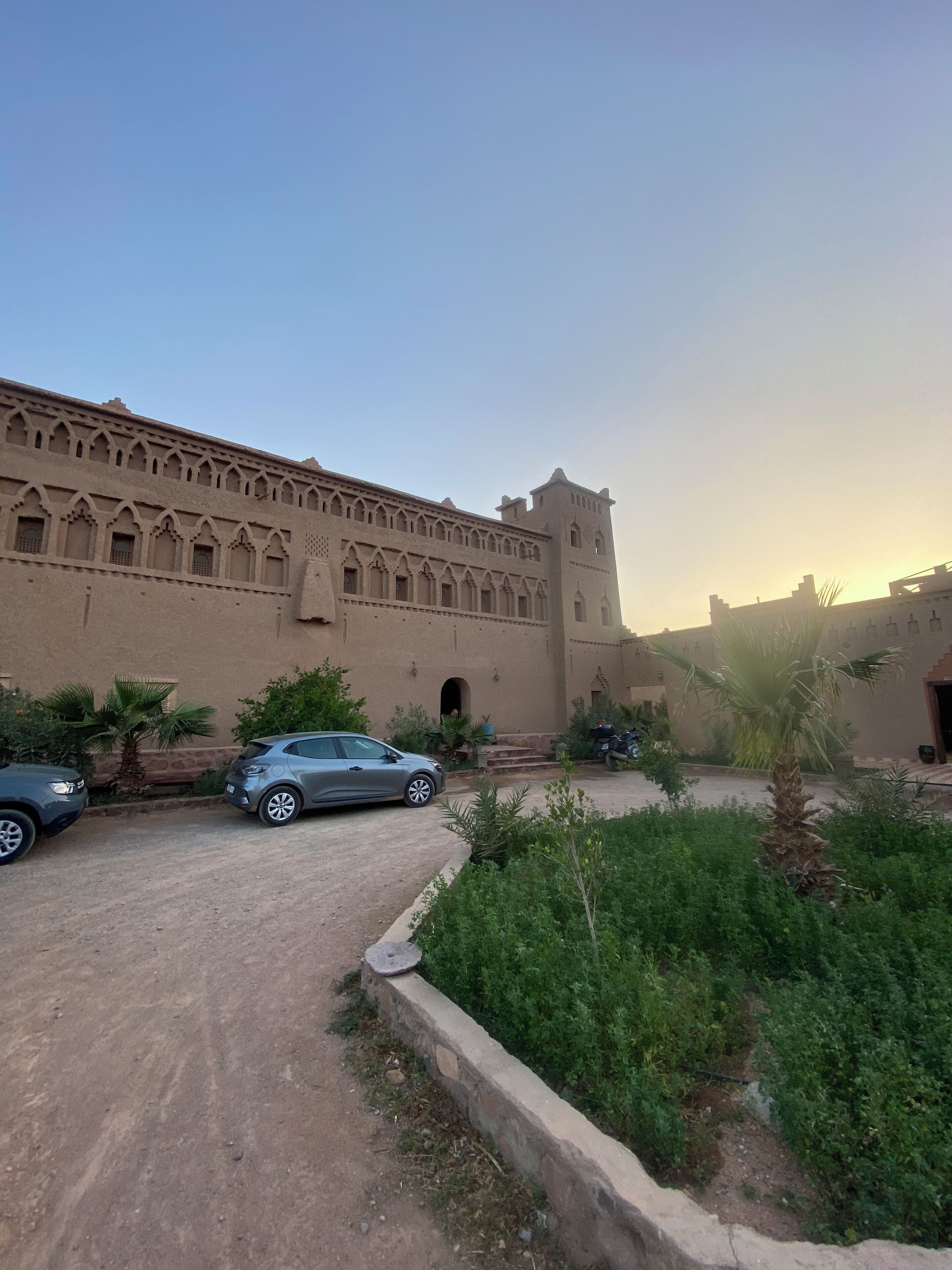 Un gran edificio con aspecto de fortaleza, que cuenta con ventanas en arco y una torre. Hay dos coches aparcados al frente, y se ve una palmera en primer plano.