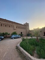 Un gran edificio con aspecto de fortaleza, que cuenta con ventanas en arco y una torre. Hay dos coches aparcados al frente, y se ve una palmera en primer plano.