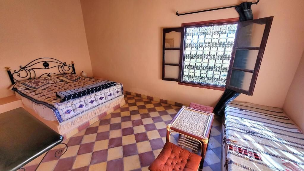 A room with a checkered floor, a bed with patterned bedding, a small table, and a window with a view.