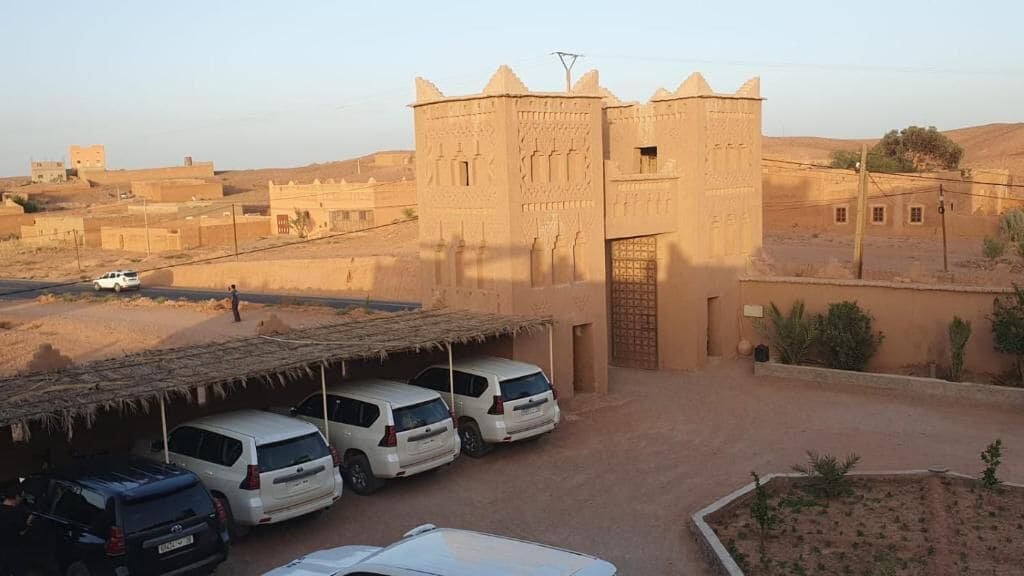 A desert landscape with a large building featuring a central tower and a covered parking area with several cars.