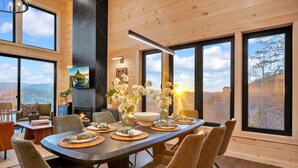 A modern dining area with a long table set for six, surrounded by large windows offering scenic views. - VIEWS, VIEWS & more VIEWS! (Sevierville)
