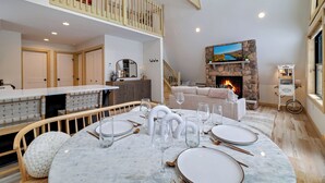 A dining area with a round table set for six, a stone fireplace, and a staircase leading to an upper level. -  Cozy Modern A-Frame with Hot tub, Fireplace, soak tub & Games (Albrightsville)