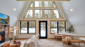 A spacious living room with a fireplace, large windows, a cozy seating area, and a dining table. -  Cozy Modern A-Frame with Hot tub, Fireplace, soak tub & Games (Albrightsville)