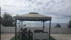A covered outdoor seating area with a table and chairs, overlooking a body of water with boats. - On the Chiaia Beach in Procida the intriguing "Whisper of the Mimant." (Procida)