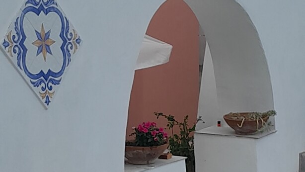 A white building with a decorative tile, a potted plant, and a small basket on a ledge. - On the Chiaia Beach in Procida the intriguing "Whisper of the Mimant." (Procida)
