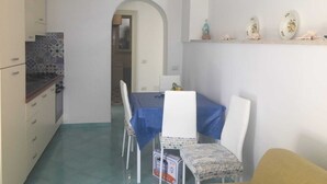 A compact kitchen with a dining area, featuring a blue tablecloth, white chairs, and a small shelf with decorative items. - On the Chiaia Beach in Procida the intriguing "Whisper of the Mimant." (Procida)