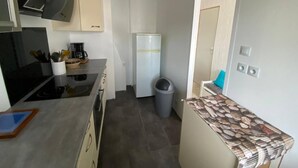 A compact kitchen with a stove, sink, and refrigerator. There is a trash bin and a small table with a patterned tablecloth. - Sea view villa, sleeps 4. (Asnelles)