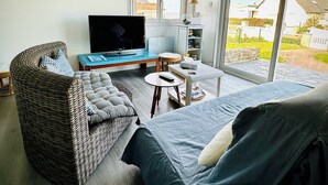 A living room with a sofa, a television, a wicker chair, a small table, and a painting of sailboats. - Sea view villa, sleeps 4. (Asnelles)