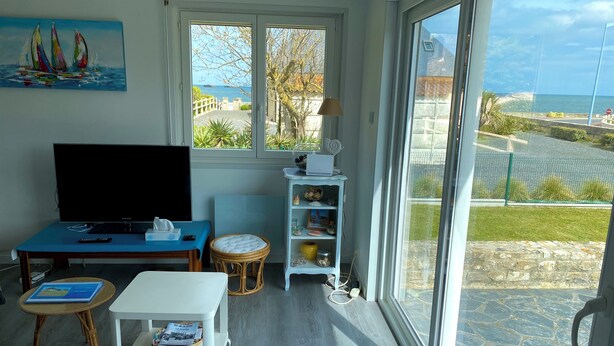 A room with a bed, a TV, a table with a remote, a wicker chair, a small table with a book, a shelf with decorative items, and a large window with a view of the sea and a building. - Sea view villa, sleeps 4. (Asnelles)