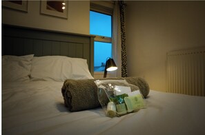 Een net opgemaakt bed met een opgerolde handdoek en toiletartikelen in een plastic zak.