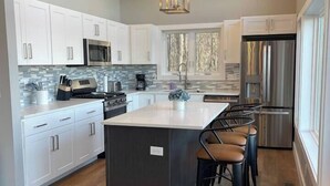A modern kitchen with white cabinets, a central island with bar stools, stainless steel appliances, and a large window providing natural light. - Lake View-5 Bedroom-Hot Tub-Kings-EV Charger, Dam View Vista-Lake View-King Beds With Luxury Linens-... (Oakland)