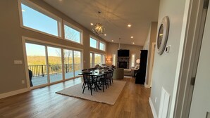 A spacious living area with a dining table, chairs, a fireplace, and large windows offering views of the outdoors. - Lake View-5 Bedroom-Hot Tub-Kings-EV Charger, Dam View Vista-Lake View-King Beds With Luxury Linens-... (Oakland)
