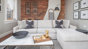 A modern living room with a sectional sofa, a coffee table with decorative items, a brick wall, and framed pictures on the wall. - Step into History: Charming Fells Point Retreat (Baltimore)