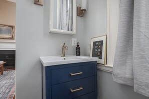 A bathroom with a white sink, a blue cabinet with gold handles, a framed picture on the wall, and a window with a curtain. - Step into History: Charming Fells Point Retreat (Baltimore)