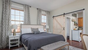 A bedroom with a large bed, bedside tables, a bench, and a view of the kitchen through an open door. - Step into History: Charming Fells Point Retreat (Baltimore)