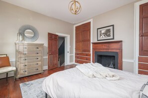 A bedroom with a large bed, a dresser, a mirror, a fireplace, and a door leading to another room. - Step into History: Charming Fells Point Retreat (Baltimore)