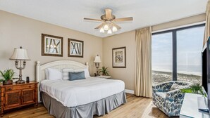 A bedroom with a bed, bedside tables, a ceiling fan, and a large window with a view of the beach.