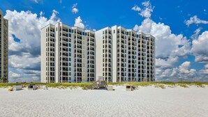 A beachfront high-rise hotel with multiple balconies, a sandy beach in the foreground, and a clear blue sky with clouds.