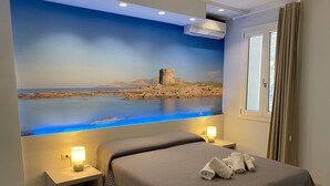 A bedroom with a large mural of a coastal landscape, a bed with a grey quilt, two bedside tables with lamps, and a window with curtains.