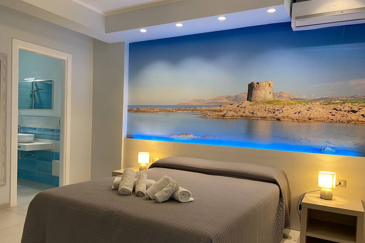 Hotelkamer met een groot bed, twee nachtkastjes met lampen, een badkamer met wastafel en spiegel, en een muurschildering van een kustlandschap.