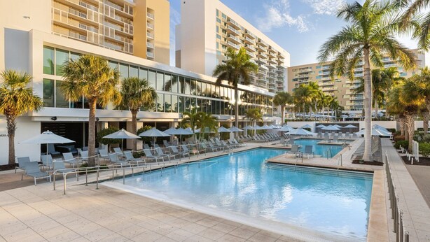 Zona de piscina de hotel con múltiples tumbonas y sombrillas, rodeada de palmeras y edificios modernos.