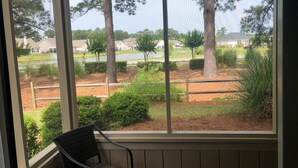 A view from a window with a chair, overlooking a landscaped area with trees, a pond, and houses in the background.