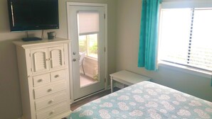A bedroom with a bed, a dresser, a television, a window with curtains, and a door leading to an outdoor area.