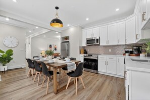 A modern kitchen with white cabinets, stainless steel appliances, and a dining area with a wooden table and black chairs. - Modern World Cup 5BR Near Downtown  Parking, Backyard & Walk to Train (Boston)