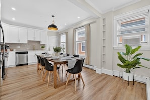 A modern kitchen with white cabinets, a dining table set for four, and a large potted plant. - Modern World Cup 5BR Near Downtown  Parking, Backyard & Walk to Train (Boston)