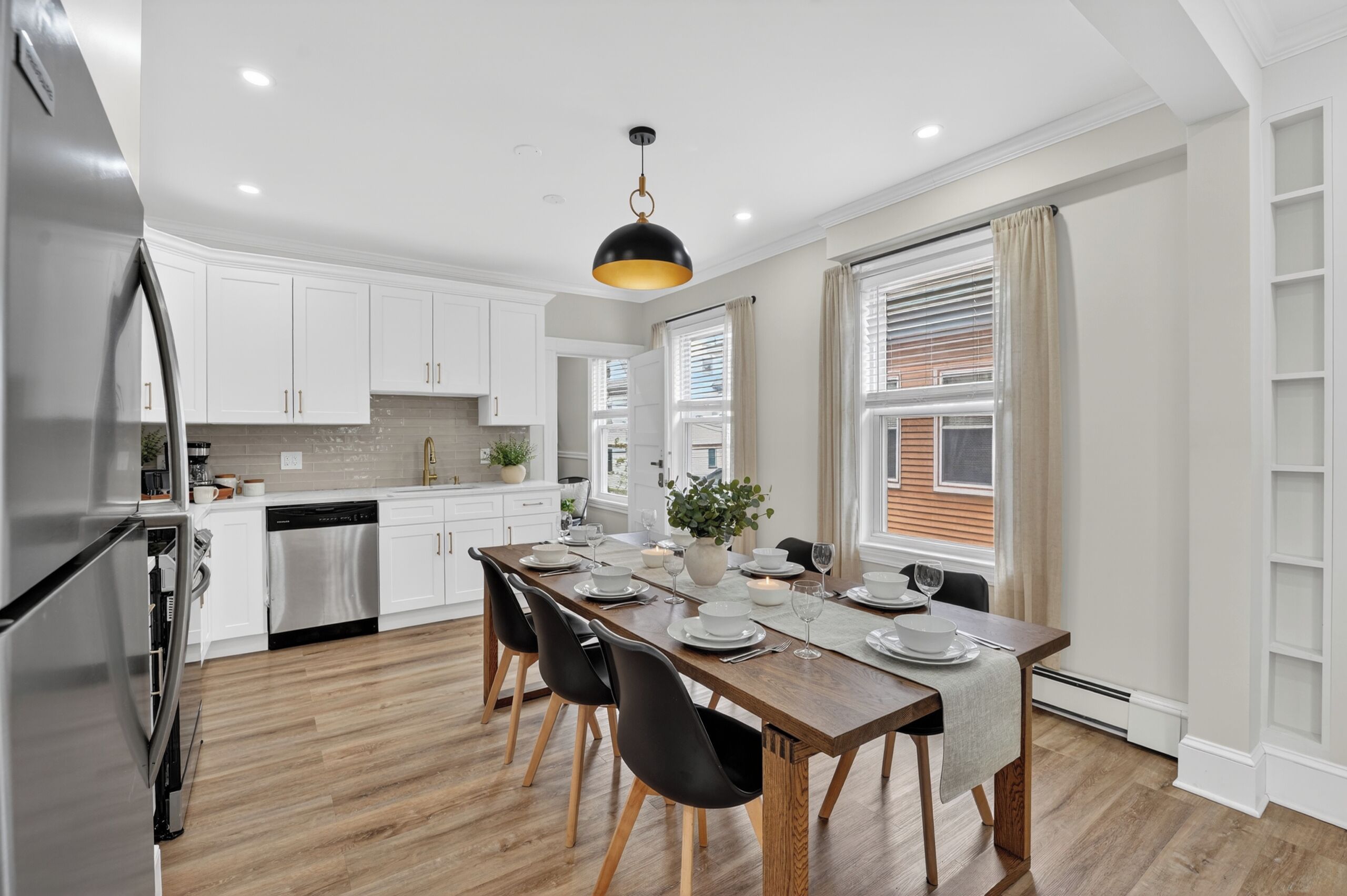 A modern kitchen with white cabinets, a stainless steel refrigerator, a wooden dining table set for four, and a pendant light.