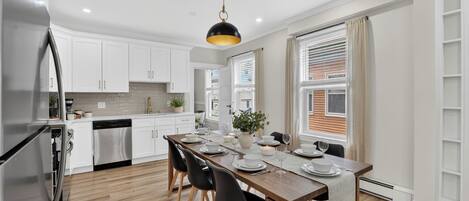 A modern kitchen with white cabinets, a stainless steel refrigerator, a wooden dining table set for four, and a pendant light.