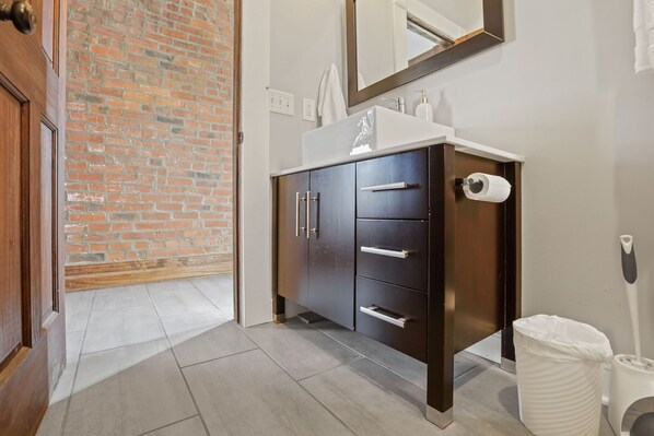 A bathroom with a brick wall, a white sink, dark cabinets, a mirror, and a trash bin.