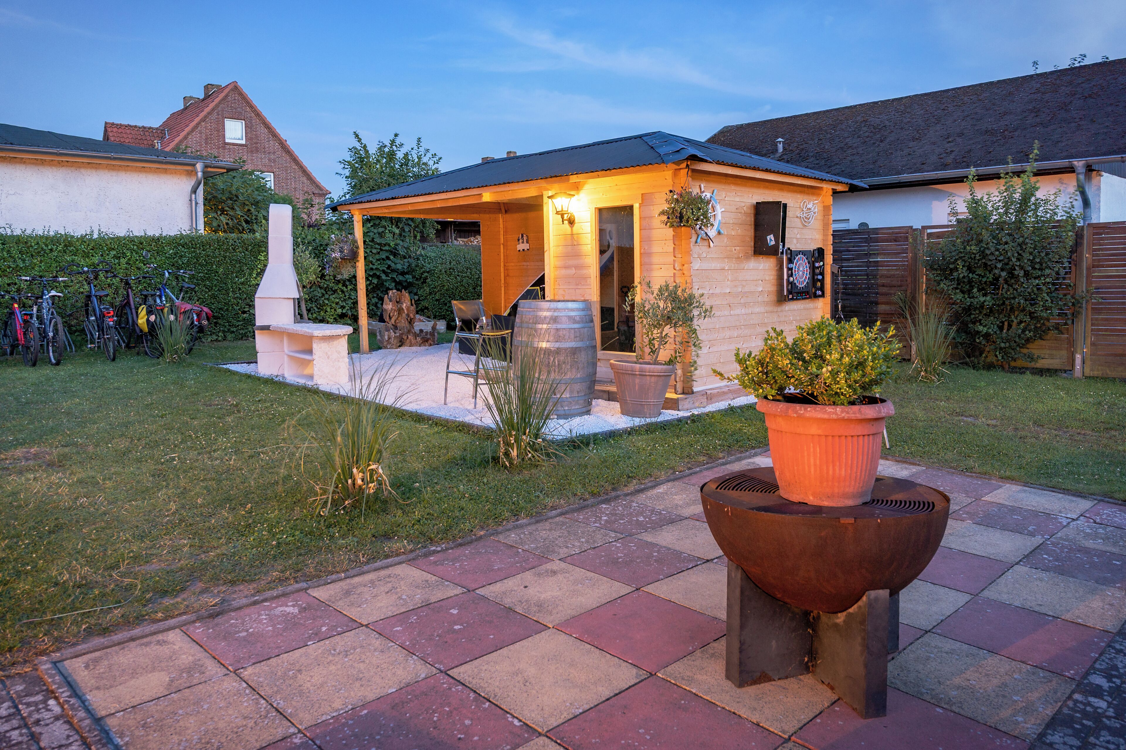Een terras met een barbecue, planten in potten en fietsen geparkeerd voor een huis met een overdekte veranda.