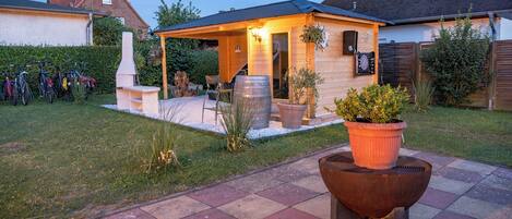 Een terras met een barbecue, planten in potten en fietsen geparkeerd voor een huis met een overdekte veranda.